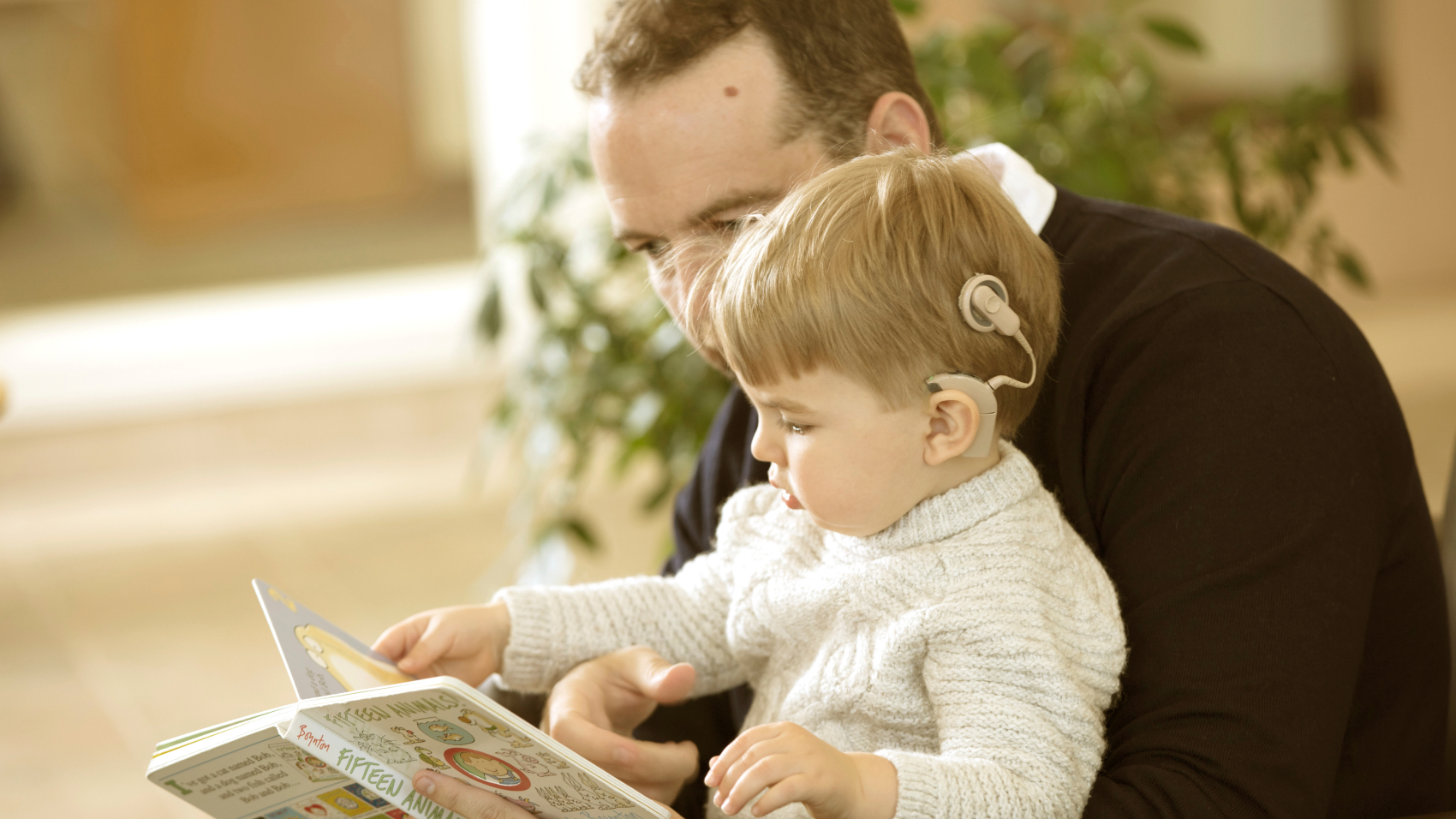 A young child sits in the lap of an adult while both are looking at a picture book.